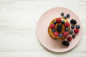 Pancakes with berries on a pink plate over white wooden surface, top view. Copy space.