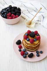 Pancakes with berries and honey on a pink plate over white wooden surface, high angle.
