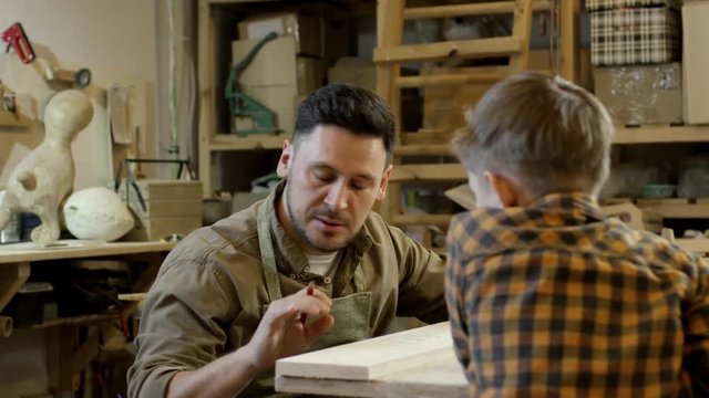 PAN of bearded male carpenter sitting at workbench and making pencil markings on wood plank while talking to little son