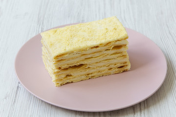Piece of Napoleon Cake on pink plate, close-up. White wooden background.