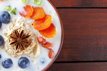 Oatmeal porridge with milk, fruits and peanuts on wooden background, healthy porridge with banana and peach slices, breakfast with fiber, calcium and vitamins, anise, dried apricots in milk