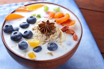 Oatmeal porridge with milk, fruits and peanuts on wooden background, healthy porridge with blueberries, banana and peach slices, breakfast with fiber, calcium and vitamins, food on blue napkin