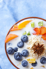 Oatmeal porridge with milk, fruits and peanuts on wooden background, healthy porridge with blueberries, banana and peach slices, breakfast with fiber, calcium and vitamins, food on blue napkin