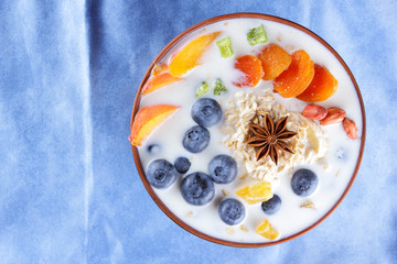 Oatmeal porridge with milk, fruits and peanuts on wooden background, healthy porridge with blueberries, banana and peach slices, breakfast with fiber, calcium and vitamins, food on blue napkin