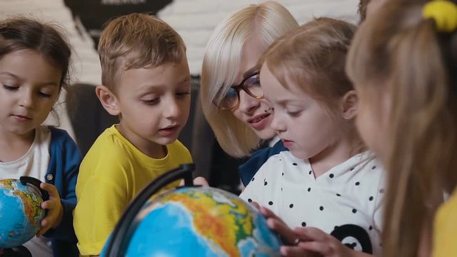 Close- up. Female young teacher and students looking the globe with a magnifying glass in the geography classroom. Cute pupils and teacher in classroom with globes at the elementary school