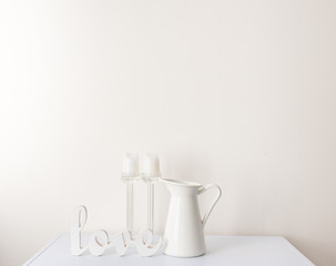 Love sign, translucent candlesticks and white jug on table against wall - white on white concept