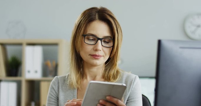 Portrait of the Caucasian good looking businesswoman sitting in her office in front of the computer screen and checking some data on the tablet device.