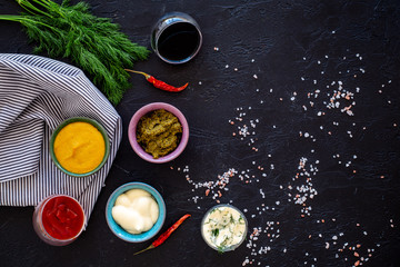 Spicy sauses. Ketchup, mayonnaise, mustard, soy sauce, barbecue sauce, pesto, mustard, sour near chili pepper and greenery on black background top view space for text
