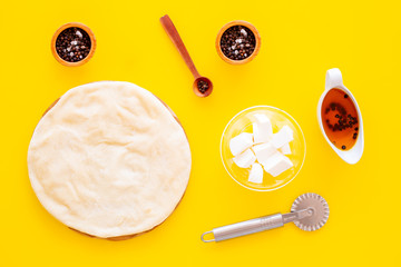 Raw rolled out pizza dough near cheese mozzarella, olive oil, spices, knife for pizza on yellow background top view mockup