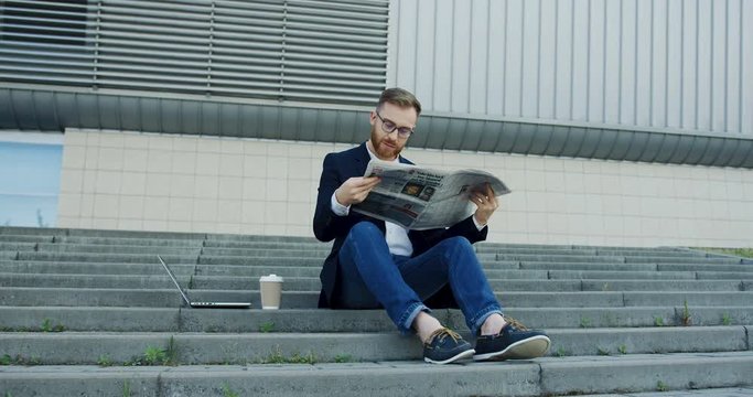 Young caucasian businessman sitting obn the city center steps and reading a newspaper while having his morning coffee to-go. Outdoor.