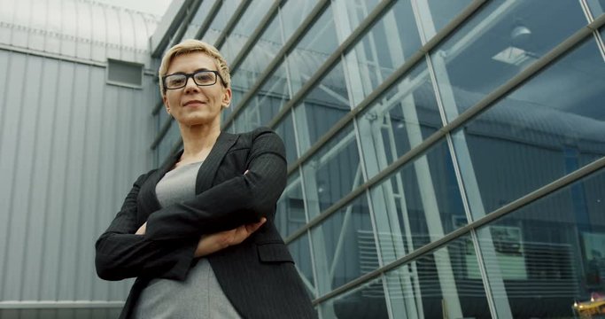 View from below on the Caucasian blonde woman in glasses posing to the camera and smiling near the big office building.