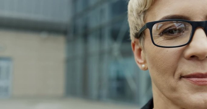 Close up of the half face in glasses of the smiled Caucasian businesswoman on the blurred background of the building.