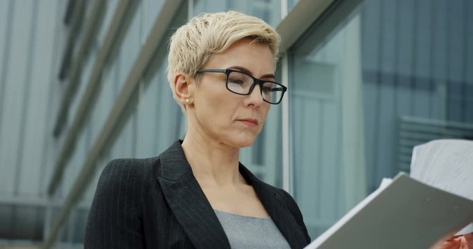 Closs up of the blond Caucasian boss woman in glasses reading and checking documents near the office center.