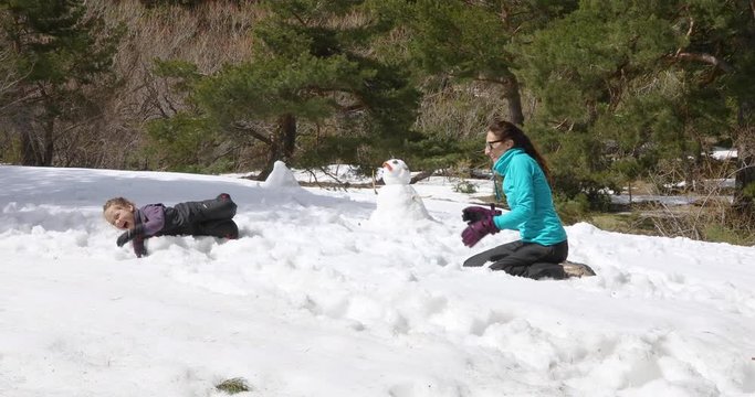 4k video in slowmotion. Woman mother throwing a snowball to four years old blonde laughing child, sitting on snow in winter mountain, next to snowman
