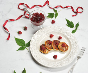 On a plate of syrniki with fresh berries of raspberries. Next to a bowl of raspberry jam. The leaves of raspberry and ribbon of crimson color complement the composition. Light background. Close-up.