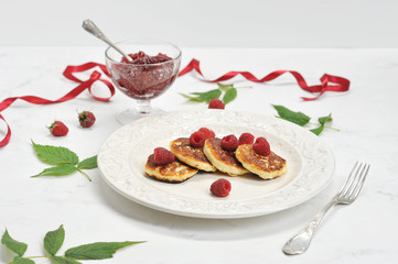On a plate of syrniki with fresh berries of raspberries. Next to a bowl of raspberry jam. The leaves of raspberry and ribbon of crimson color complement the composition. Light background. Close-up.
