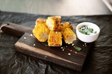 Corn cooked on the grill. Slices of corn cob on a wooden board are sprinkled with herbs and sea salt. Next cup with sauce. Black background. Close-up. 