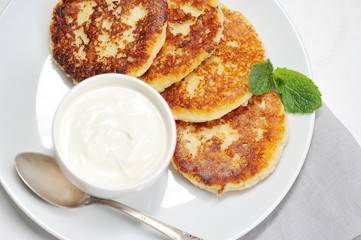 Cheesecakes on a white plate. Next to the cheesecakes is a cup of sour cream and a spoon. The cheesecakes are decorated with a leaf of fresh mint. View from above. Close-up. Macro photography.