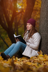 Thoughtful young female student wearing coat and knitted hat sitting in the fall park with book in hands