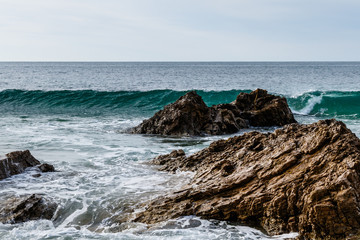 Fototapeta na wymiar Wave cresting behind rocks near the beach of Crystal Cove State Park in Laguna Beach, California. Foam from an earlier wave is in the foreground; Pacific ocean is in the background.