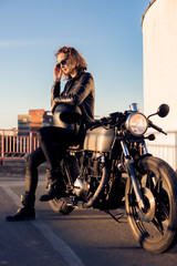 Fototapeta premium Sexy biker lady in black leather jacket sit on vintage custom caferacer motorcycle and correct sunglasses. Urban roof parking, sunset in big city. Traveling and active hipster lifestyle. Girls power.