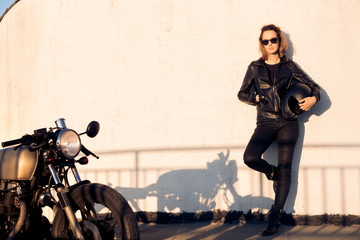 Naklejka premium Seksowna rowerzysta kobieta w czarnej skórzanej kurtce i okularach przeciwsłonecznych z rocznika caferacer zwyczajnym motocyklem blisko ściany. Miejski parking na dachu, zachód słońca w dużym mieście. Podróżowanie i aktywny styl życia hipster. Siła dziewc