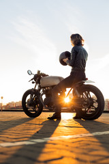 Fototapeta na wymiar Sexy biker lady in black leather jacket and full face helmet sit on vintage custom caferacer motorbike. Urban roof parking, sunset in big city. Traveling and active hipster lifestyle. Girls power.