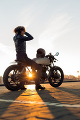 Sexy biker woman in black leather jacket sit on vintage custom caferacer motorcycle and touch her hair. Urban roof parking, sunset in big city. Traveling and active hipster lifestyle. Girls power.