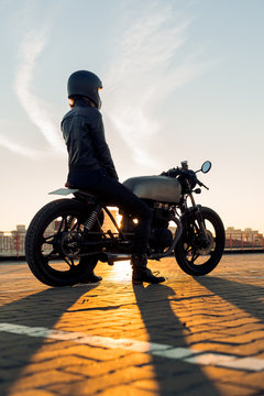 Sexy biker female in black leather jacket and full face helmet sit on vintage custom caferacer motorbike. Urban roof parking, sunset in big city. Traveling and active hipster lifestyle. Girls power.