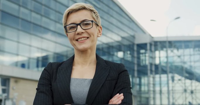 Portrait of the middle aged woman in glasses in official style smiling to the camera with hands crossed in front of her. Close up.