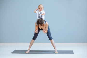 Tilt forward. European sportive mother piggybacking her toddler baby son in fitness clothing on gray background. Motherhood, healthy lifestyle concept.