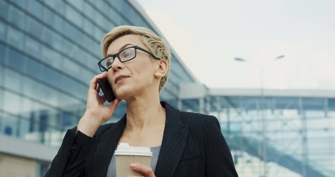 Close up odf the beautiful Caucasian woman office worker talking on the phone while drinking her morning coffee near the office building.