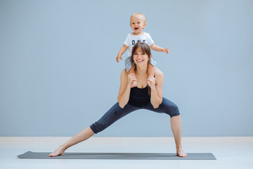 European sportive mother piggybacking her toddler baby son in fitness clothing on gray background....