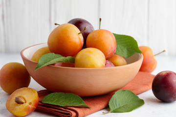Fresh colorful plums with leaves in fruit bowl on white stone background, close up view