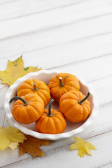 Mini orange pumpkins in a white dish and yellow leaves on white wooden background, holiday decoration