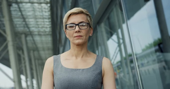 Caucasian middle aged woman in glasses standing near glass building and with serious face turning her head to the camera.