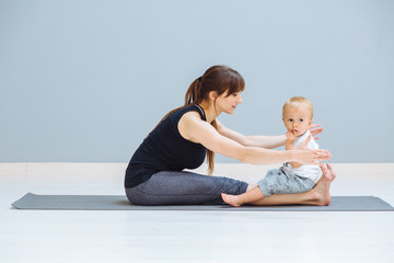 Sportive brunette mother doing stretching, workout together on yoga mat over gray wall background. Athletic and healthy motherhood. Fitness, happy maternity yoga with children concept.