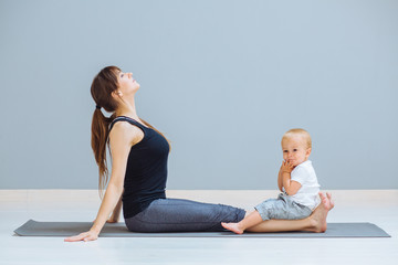 Sportive brunette mother doing stretching, workout together on yoga mat over gray wall background. Athletic and healthy motherhood. Fitness, happy maternity yoga with children concept.