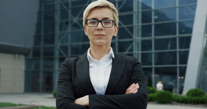 Portrait shot of the beautiful middle aged businesswoman in the glasses with hands crossed in front of her and turning her head to the camera near the big office biuilding.