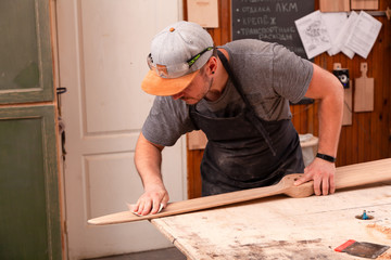 A working man in a  cap and shirt polishes the wooden block with sandpaper before painting in the workshop, in the background, tools and drilling machine.