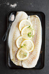 Raw cod fillets with lemon slices on a black baking tray. Top View.