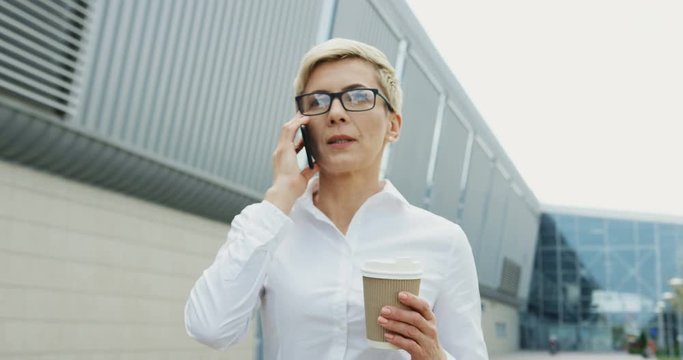 Close up of the serious blonde middle aged Caucasian businesswoman talking on the smartphone while walking with a coffee to-go in her hand to her work place.