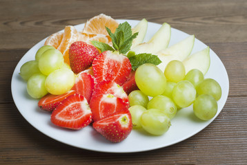 A plate with mixed fruits and sliced fruits. Delicious snack for kids or adult. Strawberries, apple, tangerine citrus and grape. Vitamine health food over wooden background. Isoalted.