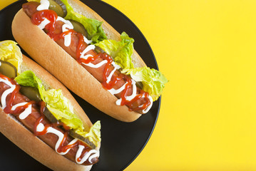 Two homemade Hot Dogs with mayonnaise, ketchup, and green lettuce leaves in black plate over yellow background. Top view. Copy space.