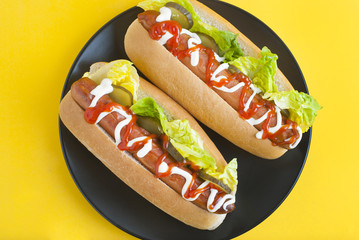 Two homemade Hot Dogs with mayonnaise, ketchup, and green lettuce leaves in black plate over yellow background. Top view.