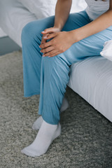 cropped shot of woman in pajamas sitting on bed and suffering from pain in knee