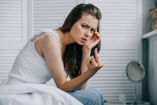 young woman with headache giving the finger and looking at camera in bedroom