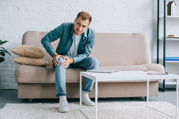 young man suffering from knee pain and looking at camera while sitting on couch