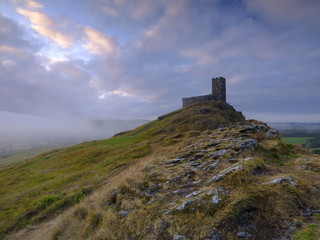 Fototapeta na wymiar Summer sunrise on Brentor showning St Michael's church atop the tor with dramatic weather clouds of showers and mist, on the western edge of the Dartmoor National Park. UK