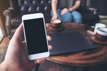 Mockup image of hand holding white mobile phone with blank black screen in cafe and woman in background